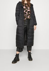 Donna che indossa un lungo cappotto trapuntato nero sopra un maglione floreale, pantaloni in pelle neri a cavallo e stivaletti neri con lacci fino alla caviglia.