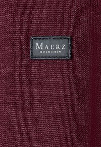 Burgunderfarbener Strickstoff mit einem rechteckigen schwarzen Etikett, auf dem "Maerz Muenchen" in weißem Text aufgenäht ist.