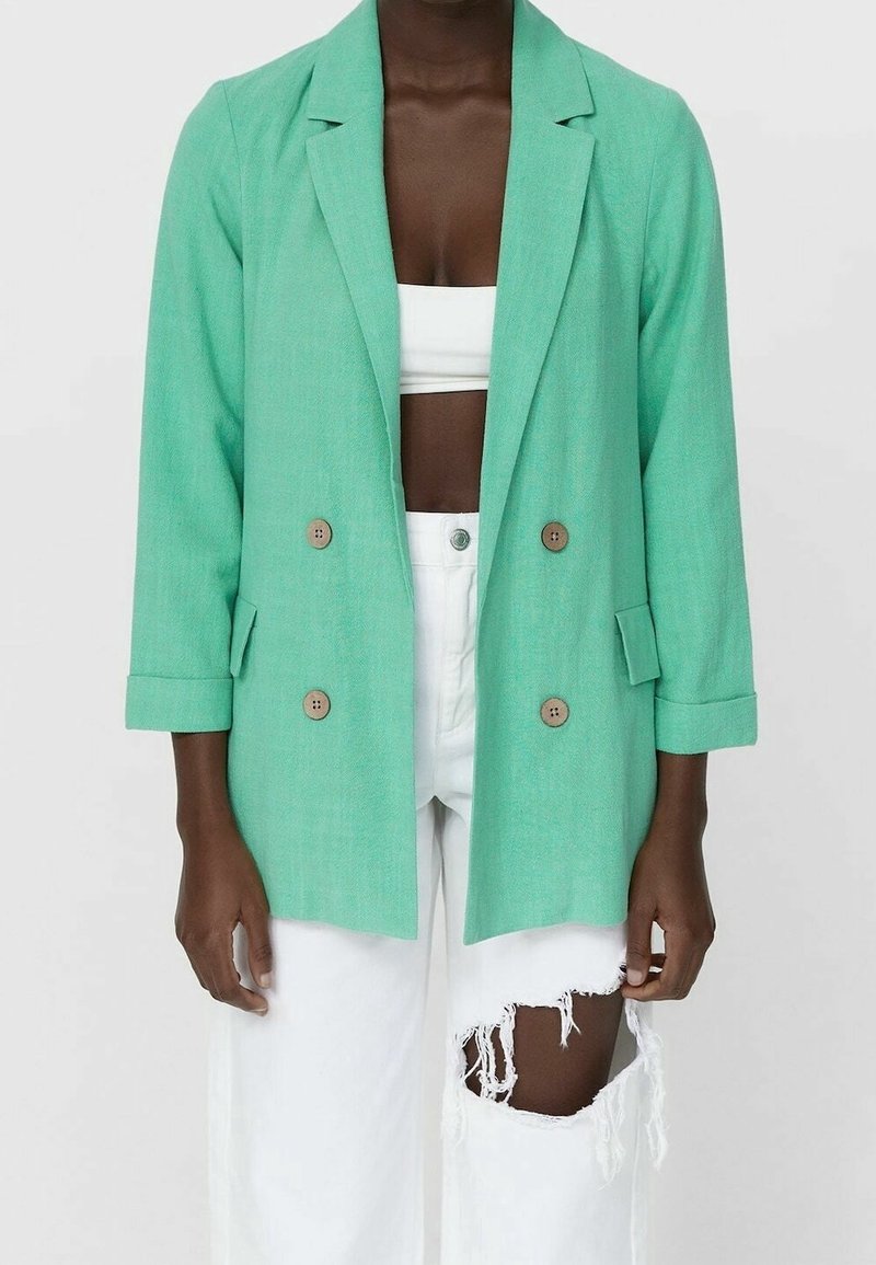 Personne portant un blazer croisé vert menthe sur un crop top blanc et un jean blanc déchiré, sur un fond uni.