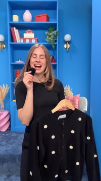 Moteris laiko juodą kardiganą su taškeliais ant pakabos ir mažą juodą pūkuotą objektą mėlyname kambaryje su lentynomis ir sieniniais šviestuvais.