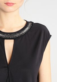 Haut noir présentant un col rond avec des embellissements texturés. Le tissu est lisse avec un léger éclat et des manches courtes.