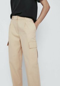 Beige cargobroek met zijzakken, gemaakt van een gladde stof. Kenmerkt zich door een relaxte pasvorm en riemlussen. De zakken hebben rechthoekige kleppen.