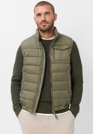 Mann mit kurzen Haaren und Bart, der eine olivgrüne gesteppte Weste über einem dunkelgrünen Pullover trägt und mit den Händen in den Taschen einer hellbeigen Hose steht.