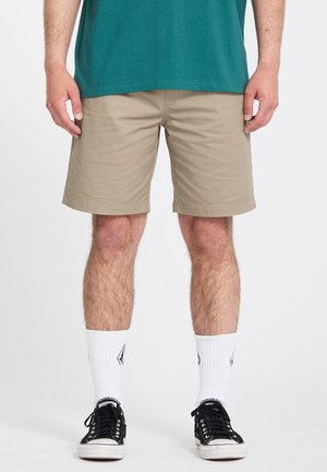 Homme portant un short beige, un t-shirt bleu sarcelle, des chaussettes blanches montantes avec des logos, et des baskets basses noires, debout contre un fond blanc.