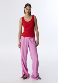 Czerwona prążkowana bluzka na ramiączkach w połączeniu z różowymi spodniami o szerokich nogawkach, z elastycznym pasem i bocznymi kieszeniami, na modelce na jednolitym tle.