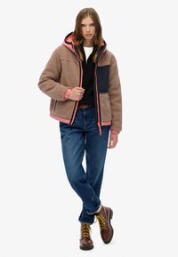 Veste en polaire marron avec panneau bleu marine, accents roses et une capuche. Portée sur un pull noir, associée à un jean bleu et des bottes marron à lacets.