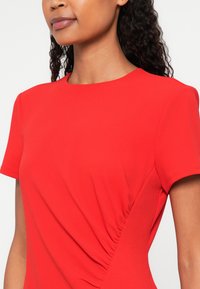 Piros, rövid ujjú blúz lekerekített nyakkivágással és oldalán redőzéssel. A sima, texturált anyag kiegészíti a testhez simuló fazont.