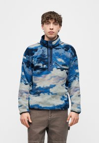 Νέος άνδρας που φοράει μπλε και γκρι φλις μπλούζα με φερμουάρ και κουμπιά, στέκεται μπροστά σε ένα απλό λευκό φόντο.