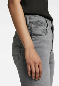 Jeans de mezclilla gris con una silueta ajustada, que presentan una etiqueta de logo negra en el bolsillo y una textura suave. Mano apoyada en el muslo.
