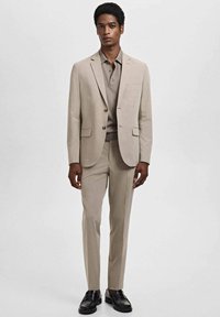 Beige kostym med en figursydd kavaj, matchande byxor och en beige polo-tröja. Har knappar på kavajen och svarta skor. Slät textur.