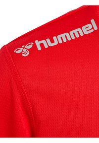Maillot de sport rouge avec un tissu texturé, des coutures proéminentes et un logo argenté représentant une abeille sur l'épaule gauche.
