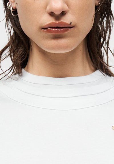 Gros plan sur la partie inférieure du visage d’une personne avec des lèvres brillantes, une tache de beauté foncée au-dessus de la lèvre, des cheveux courts et mouillés, et portant un t-shirt blanc à col rond.