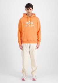 Sudadera naranja con logotipo blanco y acentos, bolsillo canguro, combinada con pantalones cargo beige claros y zapatillas multicolor.
