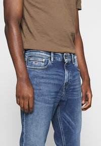 Vaqueros de mezclilla azul oscuro con un corte estándar, que presentan un logo visible en el bolsillo delantero y una ligera textura desgastada.