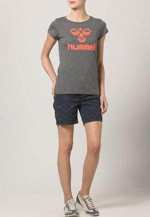 Camiseta gris de manga corta con el logo naranja de "hummel", combinada con pantalones cortos oscuros estampados y zapatillas grises. Enfoque en un atuendo casual y deportivo.