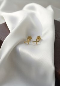 Boucles d'oreilles en plaqué or en forme de 'X', reposant sur un tissu satin blanc avec une boîte en cuir marron partiellement visible en arrière-plan.