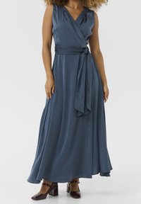 Langes, Wickelkleid aus dunkelblauem Stoff mit glatter Textur. Es hat eine gebundene Taille und einen V-Ausschnitt. Kombiniert mit dunklen, klobigen Absatzschuhen.