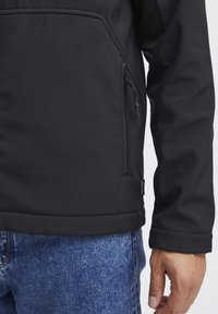 Veste softshell noire avec poche zippée, présentant une texture lisse et un design ajusté, associe à un jean en denim bleu.