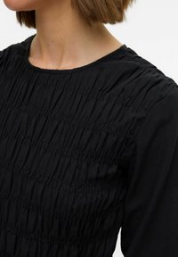 Sort stoftop med rynket tekstur, rund halsudskæring og lange ærmer. Fokus på lodrette rynkedetaljer. Glat, let materiale.