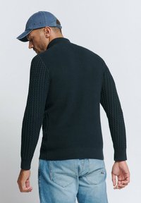 Donkere gebreide trui met een verhoogd patroon, geribbelde manchetten en zoom, gecombineerd met lichtblauwe jeans en een denim pet.