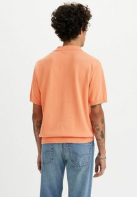 Koszulka polo z krótkim rękawem w jasnopomarańczowym kolorze, z dzianiny, z ribbrowanymi mankietami i dołem. Nosi się ją z niebieskimi dżinsami, prezentując luźny krój.