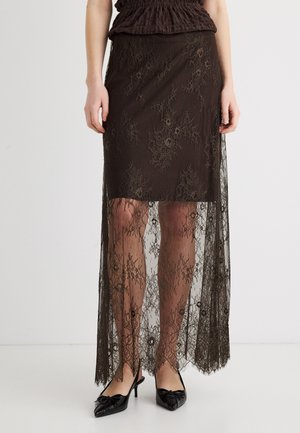 Jupe en dentelle marron avec doublure mini unie et dentelle florale transparente qui s'étend jusqu'aux chevilles, portée avec des escarpins noirs à bouts pointus et talons bas.