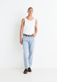White sleeveless tank top, light blue straight-leg jeans, black belt with silver buckle, brown shoes, standing against a white background.