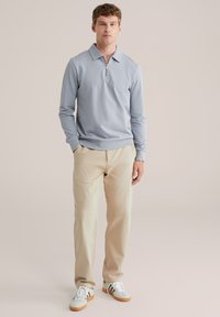 Light blue zip-up pullover, long sleeves, ribbed cuffs, paired with beige trousers and white sneakers with orange accents, plain background.