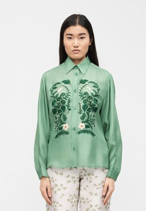 Žena v zelené košili na knoflíky s vyšívanými květinovými a ptáčími motivy, v kombinaci s bílými kalhotami s drobnými zelenými květinovými vzory.