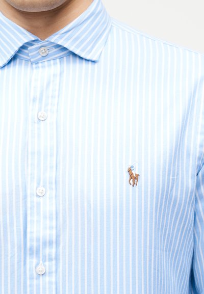 Πουκάμισο με κάθετες ρίγες σε ανοιχτό μπλε και λευκό, με κουμπιά, και μικρό κεντημένο λογότυπο παίκτη πόλο στο στήθος.