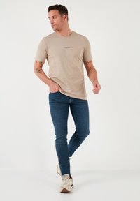 Tricou bej cu mânecă scurtă, cu un logo imprimat mic, asortat cu blugi slim-fit de culoare albastru închis și adidași de culoare deschisă.