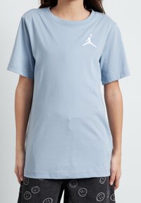 Svetle modré bavlněné tričko s krátkým rukávem, kulatým výstřihem a bílým vyšitým logem Jordan na levé straně hrudi. Hladká textura.