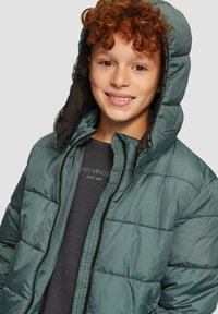  gepolsterte Jacke in dunkeltürkis, mit Kapuze, gesteppter Optik und schwarzen Akzenten. Getragen über einem dunkelgrauen Hemd mit dezentem Branding.