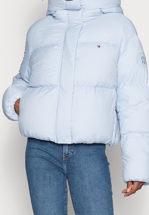 Person trägt eine hellblaue Steppjacke mit Kapuze und Tommy Hilfiger-Logo, kombiniert mit blauen Jeans. Hände in den Jackentaschen.