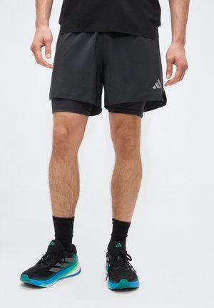 Homme portant un short de sport noir superposé, des chaussettes noires et des chaussures de course noires avec des accents turquoise et blancs, debout sur un fond blanc.