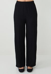 Zwarte hooggewaardeerde textuur wijd beenbroek gedragen met zwarte ballerina's, getoond van taille tot voeten tegen een effen lichte achtergrond.