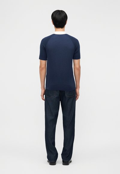 Άνδρας που στέκεται με την πλάτη προς τα εμπρός φορώντας μπλε σκούρο κοντομάνικο πουκάμισο με λευκό γιακά και σκούρα μπλε τζιν με ίσια γραμμή.