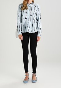 Ljusblå långärmad skjorta med knappar, prydd med ett mönster av svarta och gyllene fåglar, i kombination med svarta tajta jeans och blå platta skor med dekorationer.