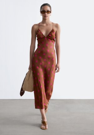 Femme portant une robe en satin marron à pois roses, sandales marron, lunettes de soleil ovales, boucles d’oreilles anneaux dorées, tenant un sac en paille beige avec un foulard.