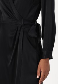 Black wrap dress made of smooth fabric featuring long sleeves, a tied belt at the waist, and a polished, elegant texture.
