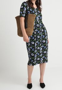 Robe noire à fleurs avec des fleurs bleues et blanches, manches courtes et silhouette ajustée. Assortis à un sac tissé marron et des chaussures noires.