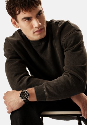 Jeune homme en sweat-shirt sombre assis, regardant sur le côté, portant une montre-bracelet noire et argentée au poignet gauche.