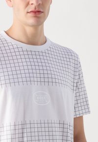 Balta marškinėliai su juodu tinklelio raštu. Apskrito kaklo iškirptė, trumpomis rankovėmis ir subtilus logotipo detalė centre. Lygi medžiagos tekstūra.