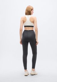 Αθλητικό crop top σε ανοιχτό κρεμ χρώμα με μαύρη λωρίδα λογότυπου· γκρι εφαρμοστά κολάν. Λεία υφή, σχέδιο με ανοικτή πλάτη και ελαφριά υλικά.