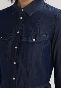 Mörkblå denimskjorta med knapp-down krage, två bröstfickor och silverknappar. Inkluderar subtila sömnadsdetaljer och en märkesetikett.