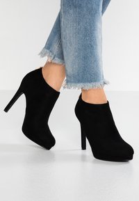 Černé semišové kotníkové boty s vysokým jehlovým podpatkem, kulatou špičkou a hladkým texturem, spárované se zdrsněnými světle modrými džíny.