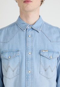 Camisa de mezclilla azul claro con botones a presión, canesú de estilo vaquero, dos bolsillos en el pecho y detalles de costura en contraste. Logo de Wrangler en el bolsillo.