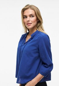 Blusa blu a maniche lunghe con vestibilità rilassata, caratterizzata da un collo senza colletto e polsini arricciati. Tessuto morbido, design minimalista.