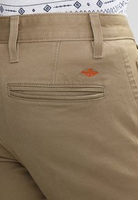 Pantaloni in cotone beige con una tasca sul retro dotata di una fessura orizzontale e un piccolo logo ricamato arancione sopra la tasca.