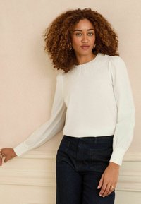 Blusa bianca a maniche lunghe con colletto in pizzo, abbinata a pantaloni in denim scuro. Il tessuto appare liscio con una leggera lucentezza.